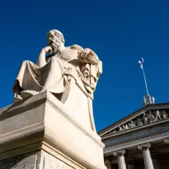 Zittend beeld van een filosoof, mogelijk Socrates, met een Griekse vlag op de achtergrond. Een eerbetoon aan de wijsheid van de godin Athene.