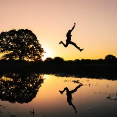 Silhouet van een persoon die springt bij zonsondergang, met een reflectie in het water. Een perfecte burst foto Samsung.