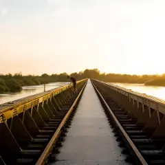 Een man leunt over een spoorbrug bij zonsondergang. De gouden gloed verlicht de rivier en de weelderige vegetatie, wat zorgt voor mooie foto locaties.