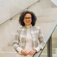 Valika Smeulders, met haar krullende haar en bril, leunt op een trapleuning. Ze draagt een stijlvolle jas en glimlacht naar de camera.