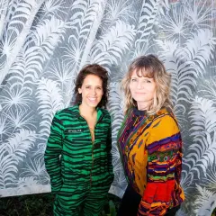 Twee vrouwen, een in een groen gestreept pak, de ander in een kleurrijke trui, staan voor een witte vitrage met palmbladeren. Geerke Catshoek lacht.
