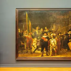 Een vrouw met blond haar en bril staat voor het beroemde nachtwacht schilderij, met een document in haar handen.