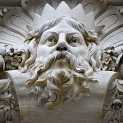 Gezicht met kroon en baard, mogelijk de vader van Zeus, omringd door bloemen en bladeren in steen.