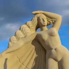 Zandstenen beeld van een **Griekse godin** met golvend haar, tegen een blauwe lucht met wolken.