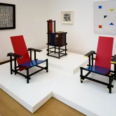 Twee iconische stoelen in rood, blauw en zwart, kenmerkend voor bauhaus kunst, staan naast een abstract schilderij en een sculptuur.