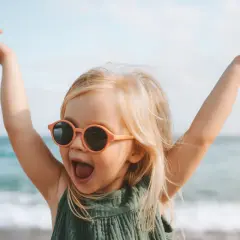 Vrolijk meisje met zonnebril op het strand, armen in de lucht. Ze lijkt wel een kleine godin, met namen als Freya of Aphrodite.