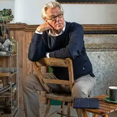 Jeroen Krabbé zit peinzend op een stoel in zijn atelier, omringd door schildersbenodigdheden.