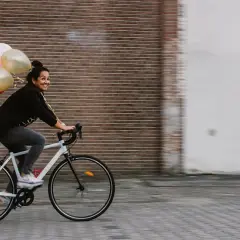 Vrolijke vrouw op een fiets met ballonnen, bewegende mensen in een stedelijke omgeving.