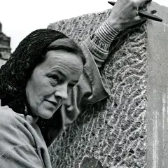 Barbara Hepworth, met een hoofddoek, bewerkt een steen met een beitel. Haar handen tonen de concentratie van een kunstenaar.