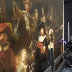 Restauratie Nachtwacht: een vrouw met mondkapje en handschoenen werkt aan het beroemde schilderij van Rembrandt.