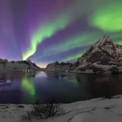 Spectaculaire foto's van het noorderlicht boven besneeuwde bergen en een bevroren meer. De groene en paarse gloed reflecteert in het water.