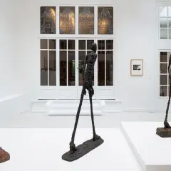 Drie slanke, bronzen beelden van l'homme qui marche staan in een witte galerie. De figuren lijken te bewegen, met hun lange benen en dunne lichamen.