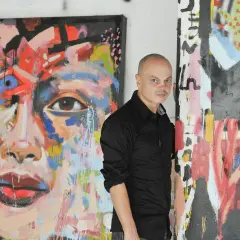 Peter Klashorst staat naast zijn kleurrijke, abstracte schilderijen. Een portret met een oog en rode lippen, en een ander werk met veel lijnen en verfspatten.