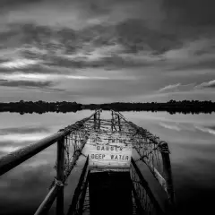Zwart-wit fotograaf legt een verlaten pier vast met een waarschuwingsbord "DANGER DEEP WATER". De weerspiegeling in het water is sereen.