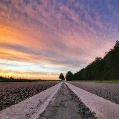 Een lege weg met witte lijnen, gefotografeerd vanuit ooghoogte perspectief. De lucht kleurt oranje en paars bij zonsondergang, met bomen aan de rechterkant.