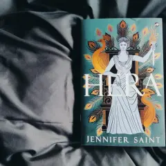 Boek "Hera" van Jennifer Saint, met de godin Hera afgebeeld met pauwenveren en een fakkel.
