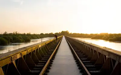 Een man leunt over een spoorbrug bij zonsondergang. De gouden gloed verlicht de rivier en de weelderige vegetatie, wat zorgt voor mooie foto locaties.