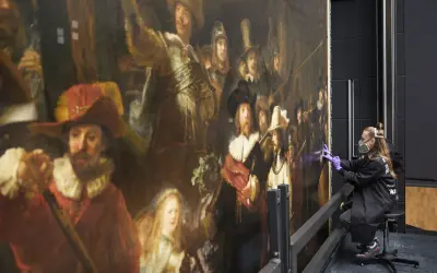 Restauratie Nachtwacht: een vrouw met mondkapje en handschoenen werkt aan het beroemde schilderij van Rembrandt.