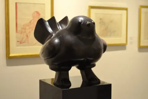 Een dikke, bronzen vogel, een typisch voorbeeld van de kunstwerken van Fernando Botero, staat op een sokkel. Op de achtergrond hangen schetsen.