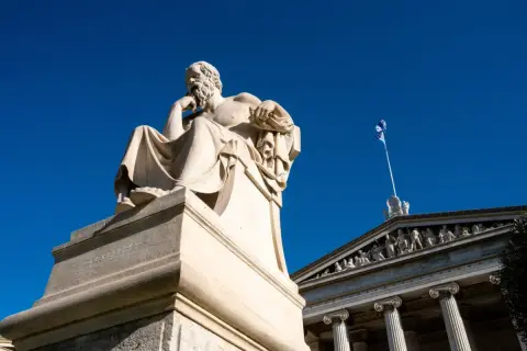 Zittend beeld van een filosoof, mogelijk Socrates, met een Griekse vlag op de achtergrond. Een eerbetoon aan de wijsheid van de godin Athene.