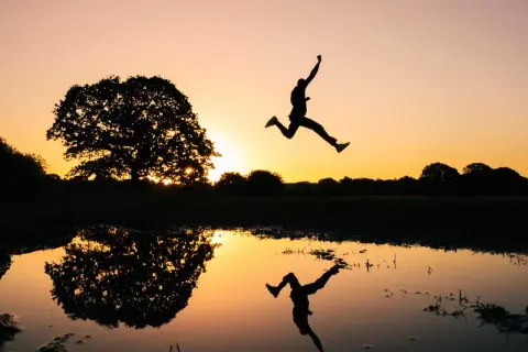 Silhouet van een persoon die springt bij zonsondergang, met een reflectie in het water. Een perfecte burst foto Samsung.