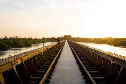 Een man leunt over een spoorbrug bij zonsondergang. De gouden gloed verlicht de rivier en de weelderige vegetatie, wat zorgt voor mooie foto locaties.