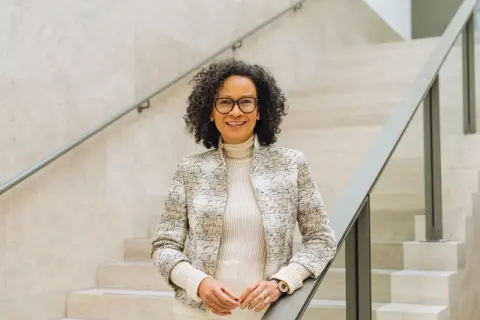 Valika Smeulders, met haar krullende haar en bril, leunt op een trapleuning. Ze draagt een stijlvolle jas en glimlacht naar de camera.