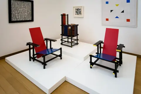 Twee iconische stoelen in rood, blauw en zwart, kenmerkend voor bauhaus kunst, staan naast een abstract schilderij en een sculptuur.