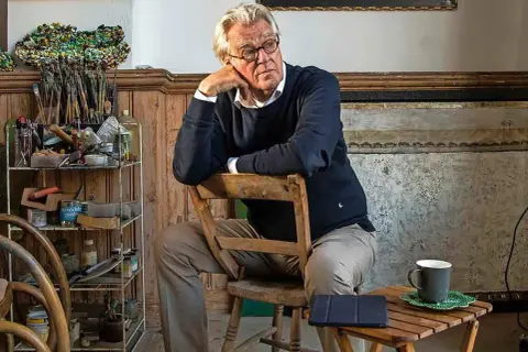 Jeroen Krabbé zit peinzend op een stoel in zijn atelier, omringd door schildersbenodigdheden.