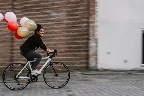 Vrolijke vrouw op een fiets met ballonnen, bewegende mensen in een stedelijke omgeving.