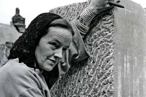 Barbara Hepworth, met een hoofddoek, bewerkt een steen met een beitel. Haar handen tonen de concentratie van een kunstenaar.