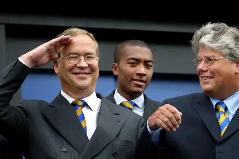 Pim de Lange salueert met een brede glimlach, naast twee mannen in pak.