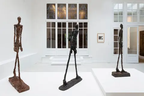 Drie slanke, bronzen beelden van l'homme qui marche staan in een witte galerie. De figuren lijken te bewegen, met hun lange benen en dunne lichamen.