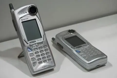 Twee zilverkleurige mobieltjes, waaronder de Kyocera VP-210, de eerste telefoon met camera.