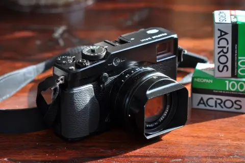 Zwarte Fujifilm X-E1 camera met lenskap en filmrolletjes Acros 100 op een houten tafel.