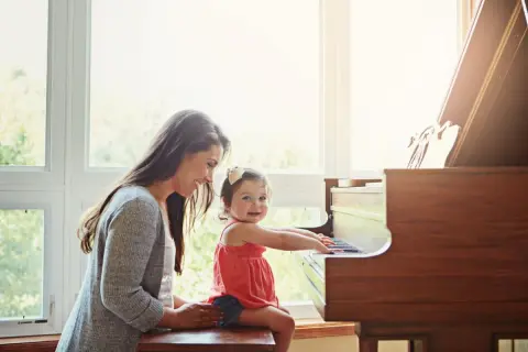 Een moeder helpt haar dochtertje met het spelen op de piano. Het kleine meisje zit op de pianokruk en lacht vrolijk terwijl ze de toetsen aanraakt.