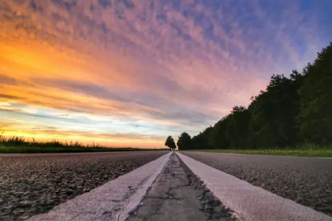 Een lege weg met witte lijnen, gefotografeerd vanuit ooghoogte perspectief. De lucht kleurt oranje en paars bij zonsondergang, met bomen aan de rechterkant.