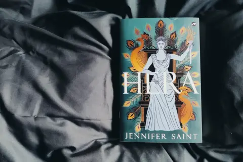 Boek "Hera" van Jennifer Saint, met de godin Hera afgebeeld met pauwenveren en een fakkel.