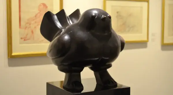 Een dikke, bronzen vogel, een typisch voorbeeld van de kunstwerken van Fernando Botero, staat op een sokkel. Op de achtergrond hangen schetsen.