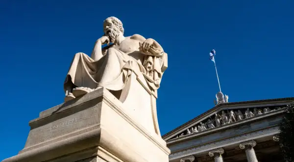 Zittend beeld van een filosoof, mogelijk Socrates, met een Griekse vlag op de achtergrond. Een eerbetoon aan de wijsheid van de godin Athene.