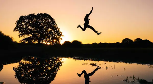 Silhouet van een persoon die springt bij zonsondergang, met een reflectie in het water. Een perfecte burst foto Samsung.