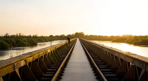 Een man leunt over een spoorbrug bij zonsondergang. De gouden gloed verlicht de rivier en de weelderige vegetatie, wat zorgt voor mooie foto locaties.