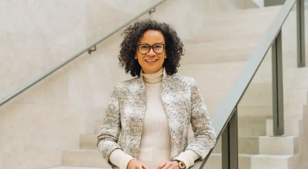 Valika Smeulders, met haar krullende haar en bril, leunt op een trapleuning. Ze draagt een stijlvolle jas en glimlacht naar de camera.