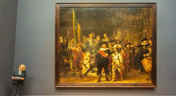 Een vrouw met blond haar en bril staat voor het beroemde nachtwacht schilderij, met een document in haar handen.