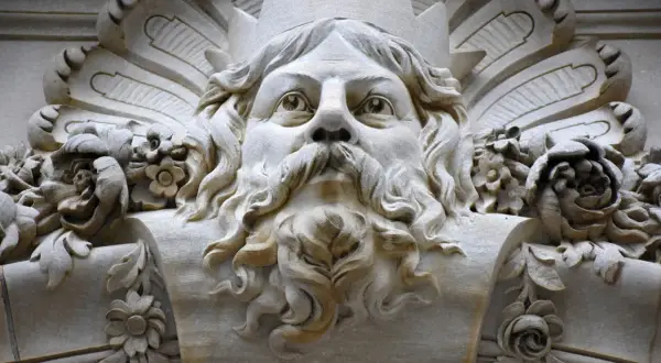 Gezicht met kroon en baard, mogelijk de vader van Zeus, omringd door bloemen en bladeren in steen.