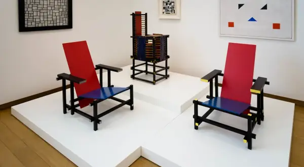 Twee iconische stoelen in rood, blauw en zwart, kenmerkend voor bauhaus kunst, staan naast een abstract schilderij en een sculptuur.