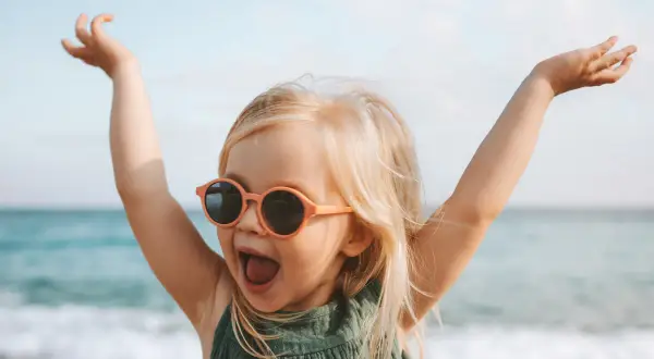 Vrolijk meisje met zonnebril op het strand, armen in de lucht. Ze lijkt wel een kleine godin, met namen als Freya of Aphrodite.