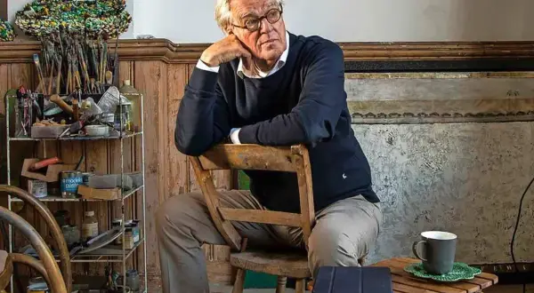 Jeroen Krabbé zit peinzend op een stoel in zijn atelier, omringd door schildersbenodigdheden.