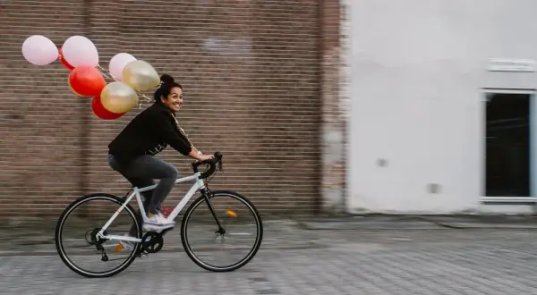 Vrolijke vrouw op een fiets met ballonnen, bewegende mensen in een stedelijke omgeving.