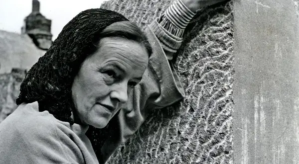 Barbara Hepworth, met een hoofddoek, bewerkt een steen met een beitel. Haar handen tonen de concentratie van een kunstenaar.
